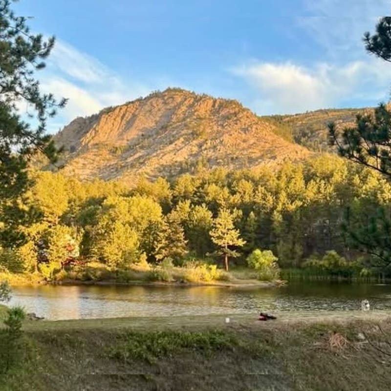 A grassy shoreline and calm lake sit in the foreground, leading to a dense forest and a large, sunlit rocky mountain under a blue sky.