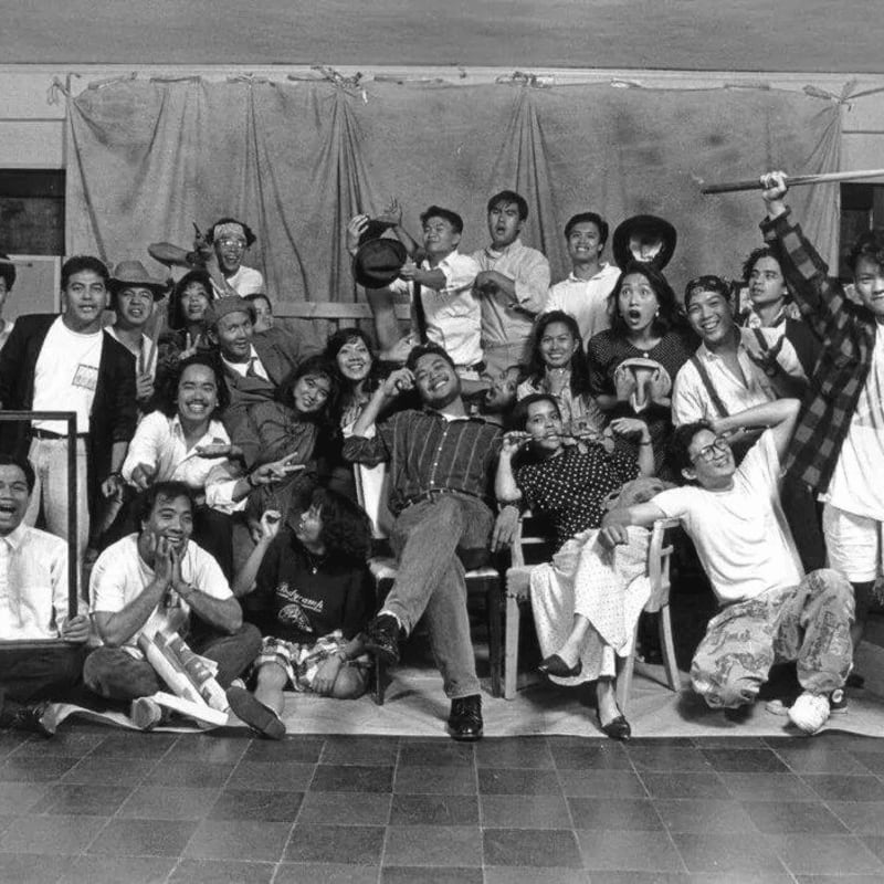 A large group of smiling people pose together in a crowded indoor setting against a backdrop of draped curtains.