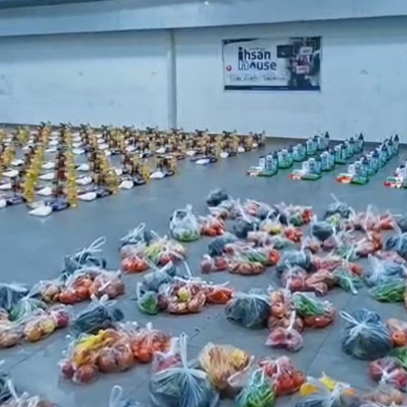 Numerous bags of produce are scattered across the foreground of a large room, with organized rows of food packages and a sign for "Ihsan House" in the background.