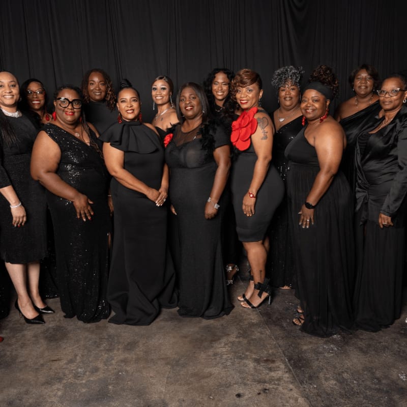 A group of women dressed in elegant black attire pose together against a backdrop of dark curtains.