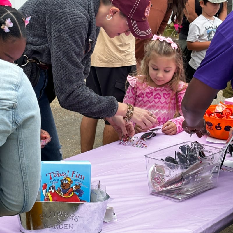 A woman and a young girl in a pink heart-patterned shirt look at small items on a purple table, with other people standing in the background.