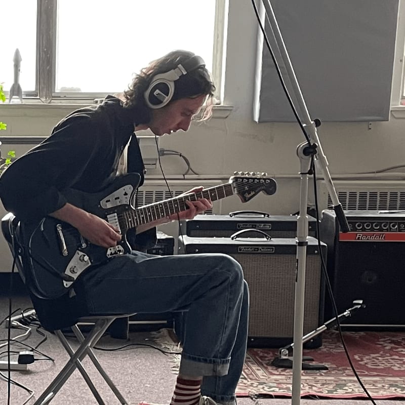 A man wearing headphones plays an electric guitar while sitting in a room with amplifiers and a microphone stand.