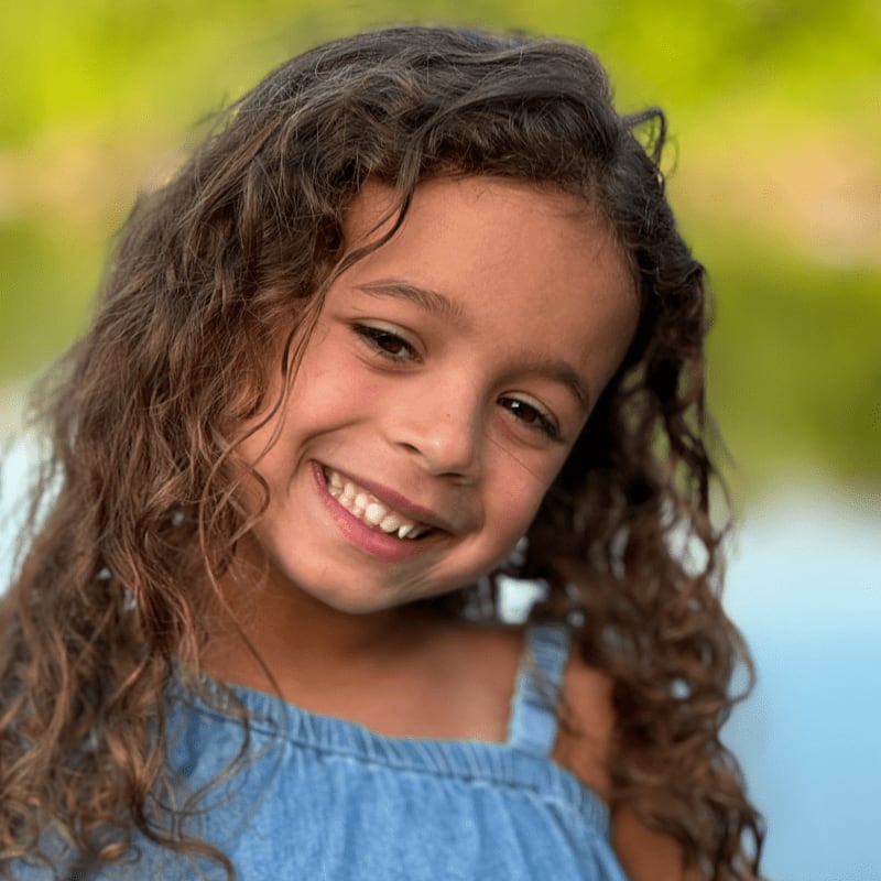 A smiling young girl with curly brown hair and a blue dress stands against a blurred green and blue background.