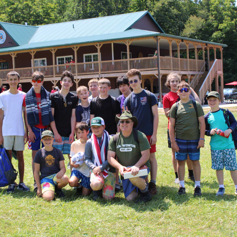 A group of boys and a man pose for a photo on a grassy lawn in front of a large, two-story wooden lodge with a green roof.