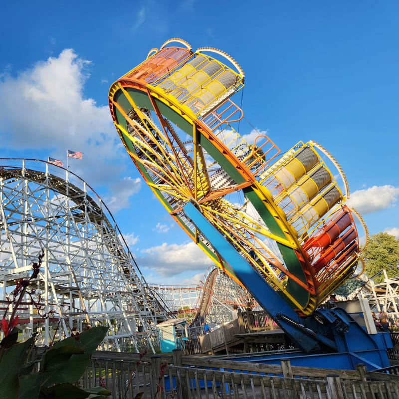 A colorful, rotating amusement park ride stands in the foreground against a backdrop of a white wooden roller coaster and a bright blue sky.