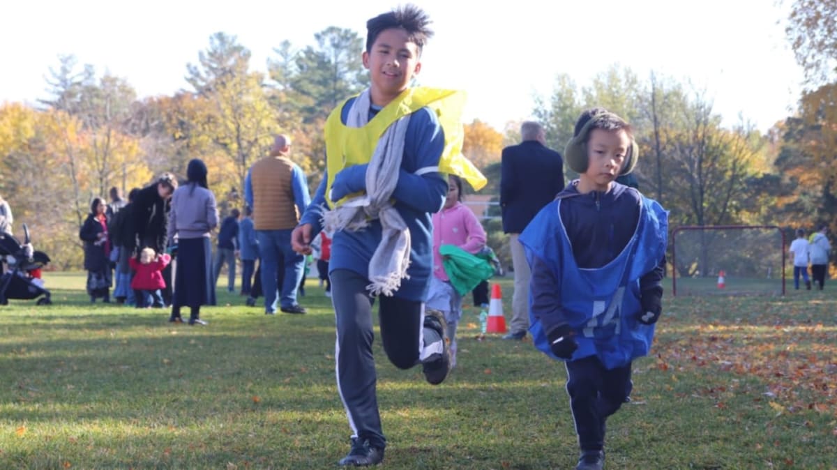 Two boys wearing colorful pinnies run across a grassy field with a crowd of people and autumn trees in the background.