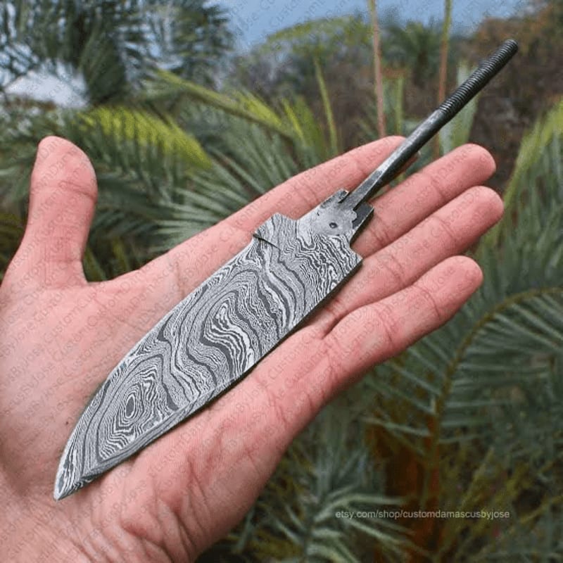 A hand holds a Damascus steel knife blade with a swirling pattern, set against a blurred background of palm fronds.
