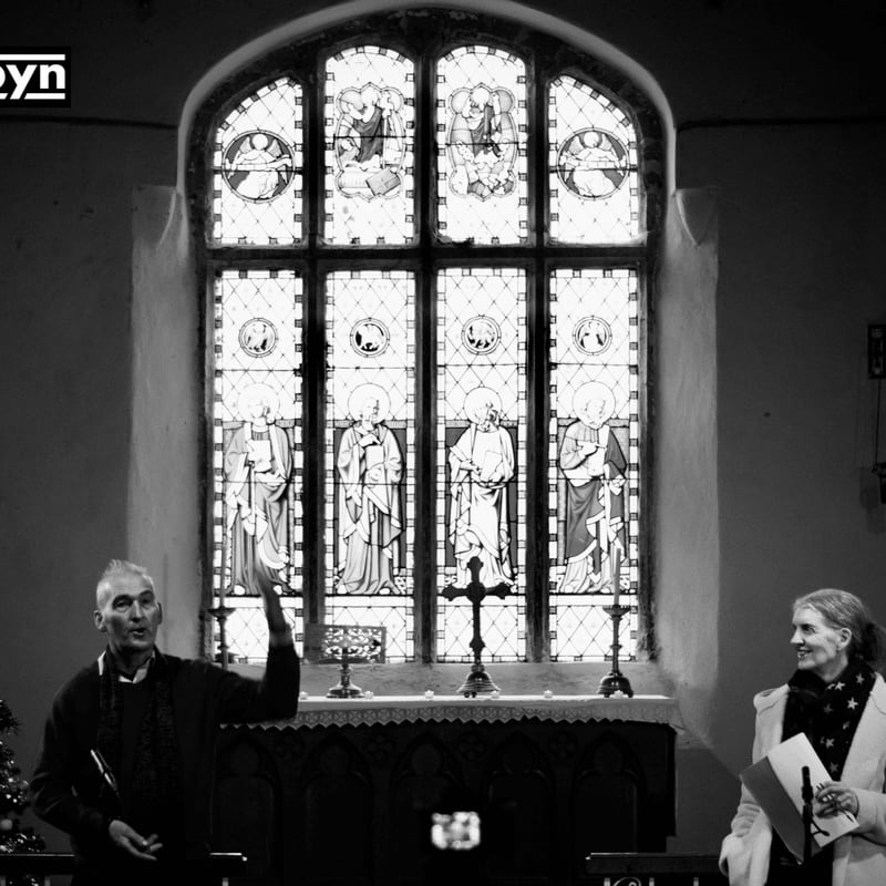 Two people stand in the foreground of a church, facing each other in front of a large stained-glass window in the background.