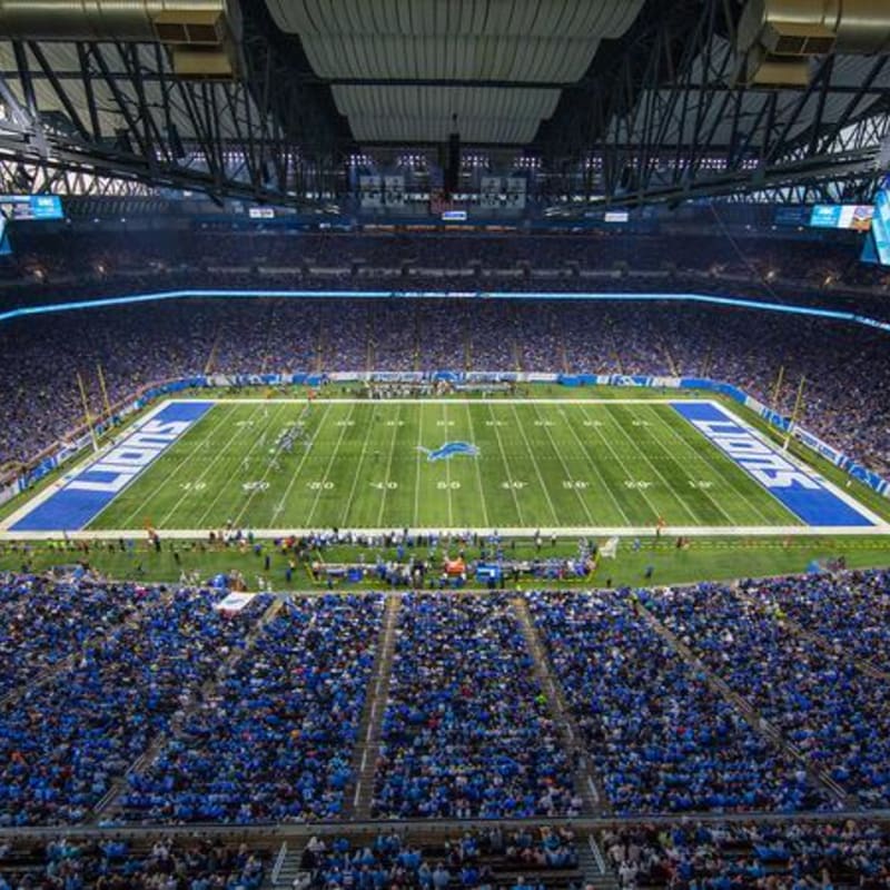 A crowded stadium of fans in blue fills the foreground, overlooking a green football field with "LIONS" end zones under a massive indoor roof.