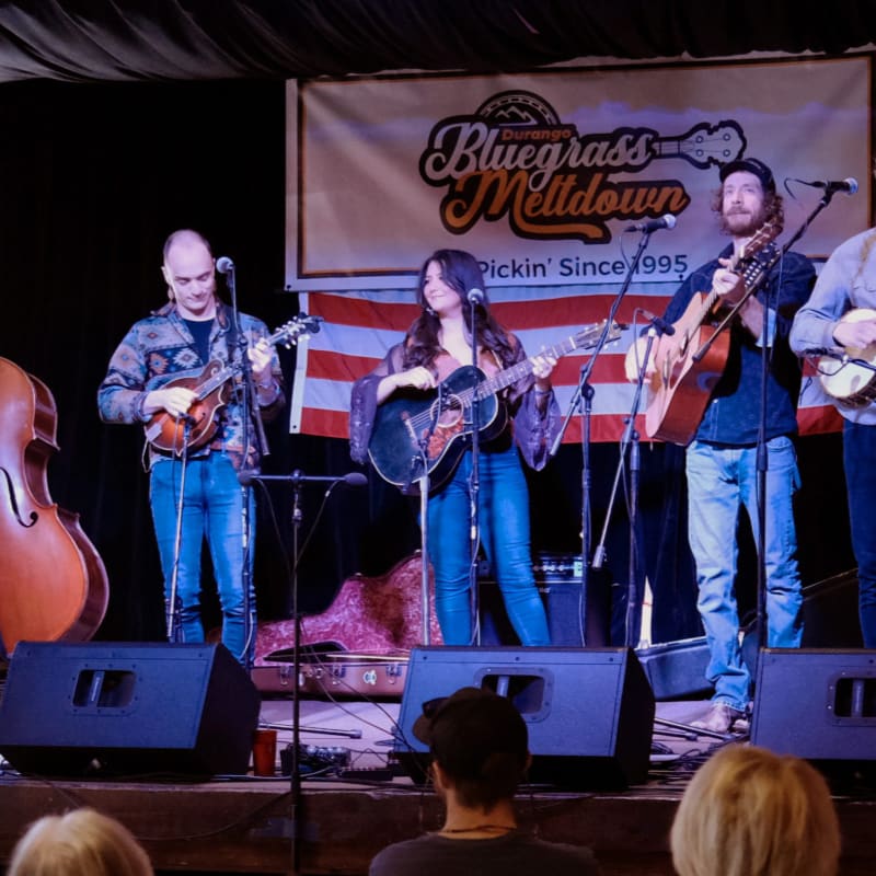 A bluegrass band performs on a stage with a "Durango Bluegrass Meltdown" banner in the background, while audience members watch from the foreground.