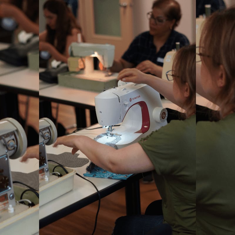 A woman in a green shirt uses a white sewing machine in the foreground, while other people work at tables in the blurred background.