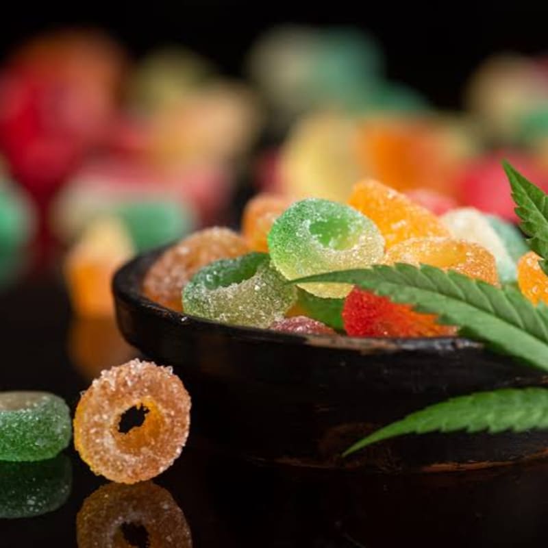 Colorful sugar-coated gummy rings fill a dark bowl with a cannabis leaf, set against a blurred background of more candies.