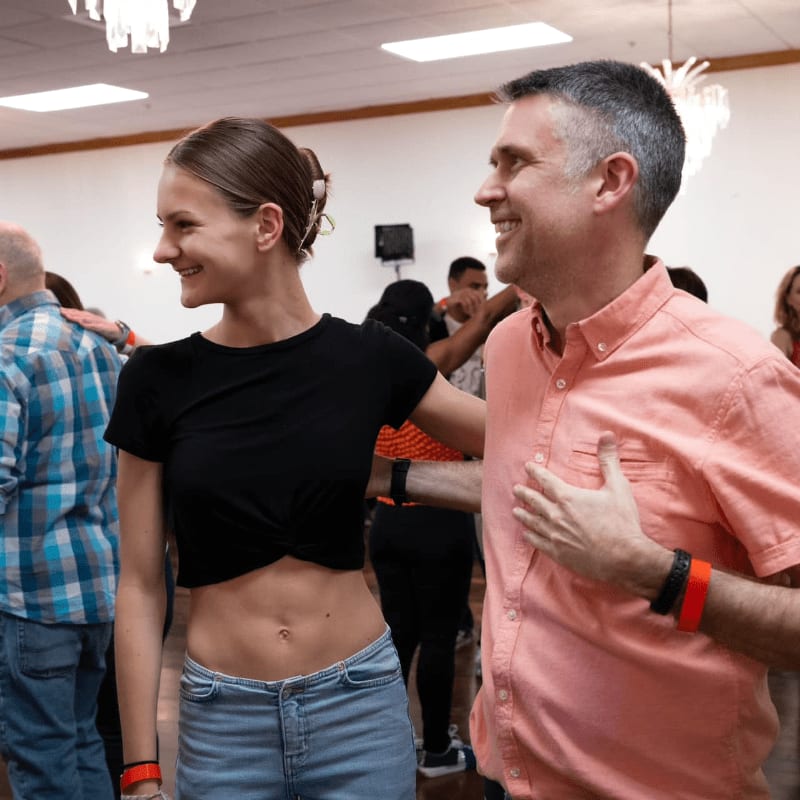 A smiling woman in a black crop top and a man in a peach shirt stand together in the foreground of a brightly lit room filled with other people.