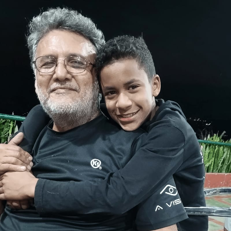 A young boy hugs an older man with glasses and a grey beard against a dark night background with greenery.