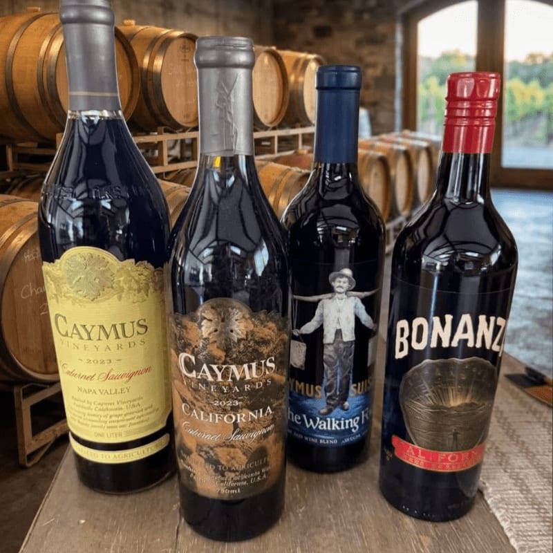 Four bottles of red wine stand in the foreground against a background of wooden wine barrels in a cellar.