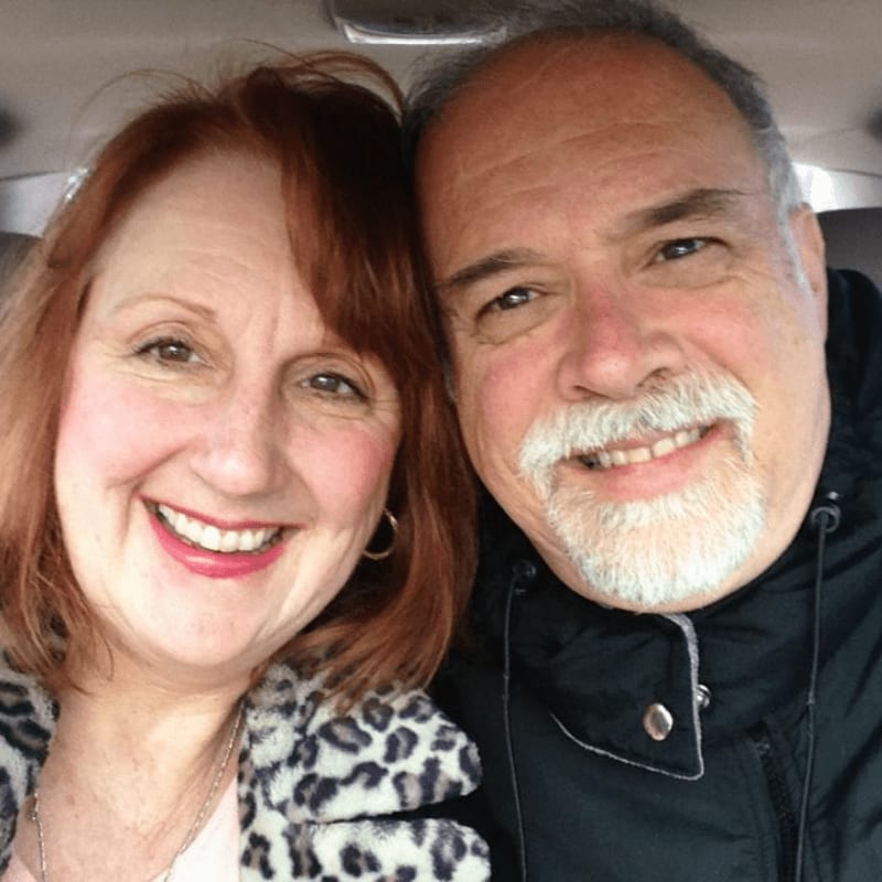 A smiling woman with red hair and a man with a grey beard pose closely together inside a car.
