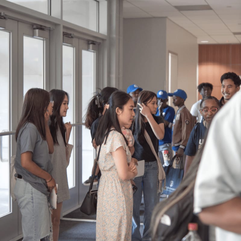 A group of young people stand and talk in a brightly lit hallway near a set of glass doors.