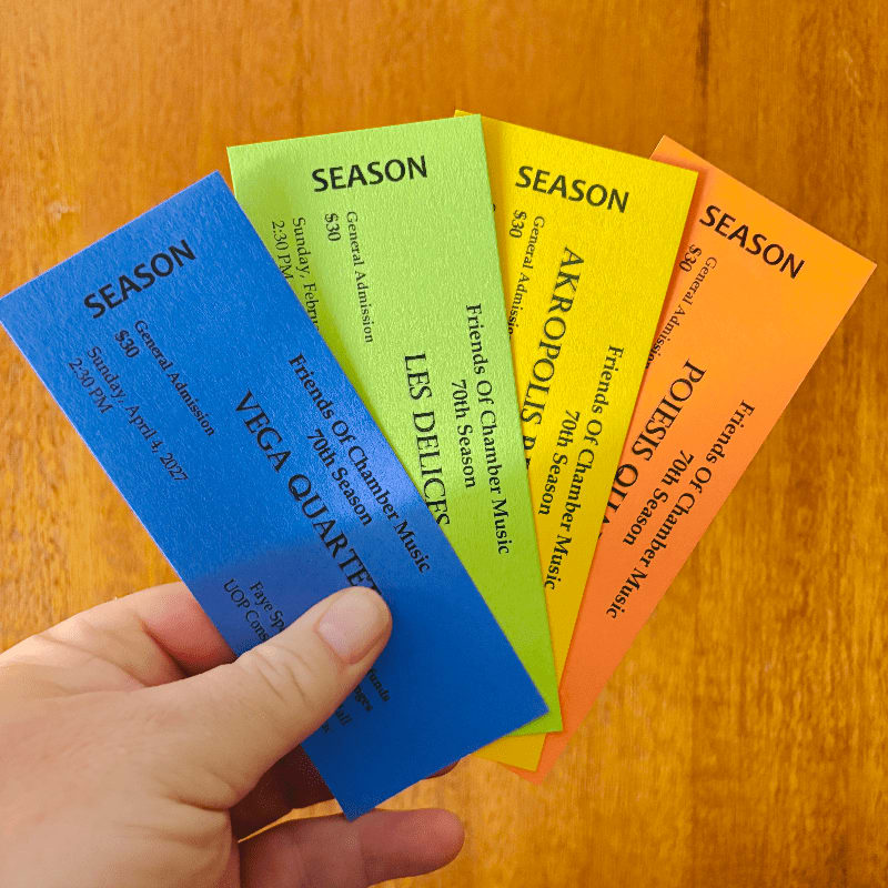 A hand holds four colorful concert tickets in the foreground against a wooden table background.