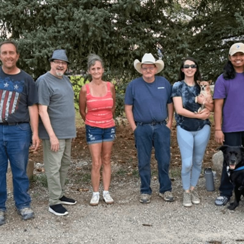 Six people and two dogs stand together on a gravel path in front of a dense backdrop of green trees.