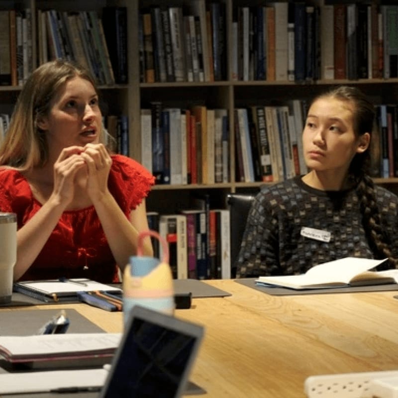 Two women sit at a wooden table in the foreground, with bookshelves filled with books serving as the background.