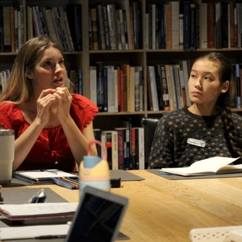 Two women sit at a wooden table with notebooks and a laptop in the foreground, set against a background of bookshelves filled with books.