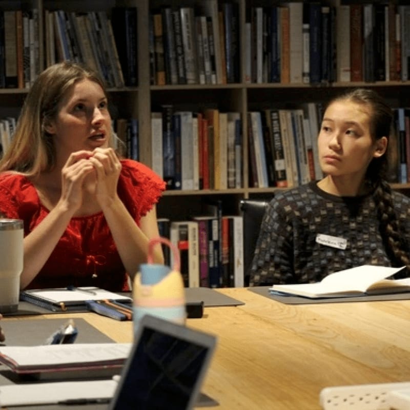 Two women sit at a wooden table with notebooks and a laptop in the foreground, set against a background of bookshelves filled with books.