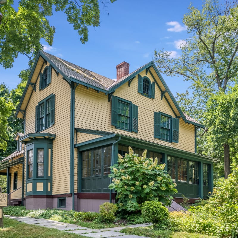 A yellow Victorian house with green trim and shutters stands amidst lush green trees and foliage.