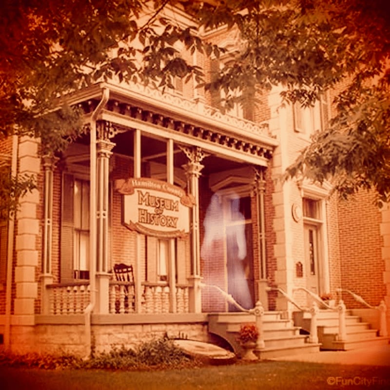 A translucent ghost stands on the porch of a brick Museum of History, framed by overhanging tree branches.