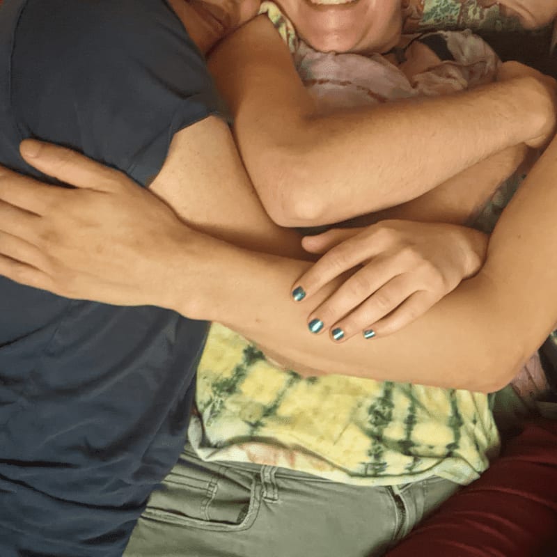 Two people embrace in the foreground, with one wearing a blue shirt and the other a tie-dye shirt, while a colorful, patterned fabric is visible in the background.