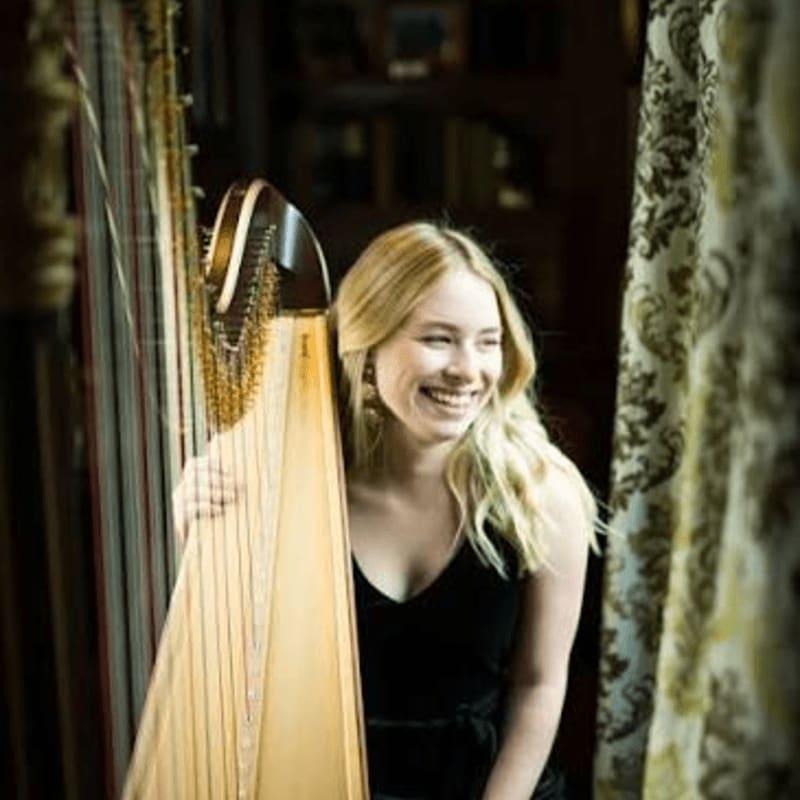 A smiling blonde woman in a black dress stands beside a harp in a dimly lit room with patterned curtains.