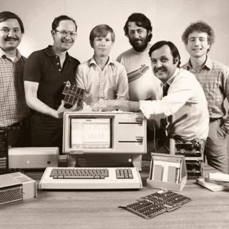 Six men stand behind a table featuring early computer hardware and a monitor in a plain indoor setting.