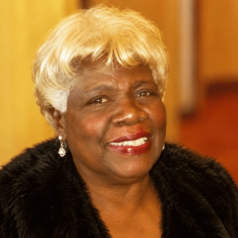 A smiling woman with blonde hair and a black faux-fur coat is positioned in the foreground against a blurred, warm-toned background.