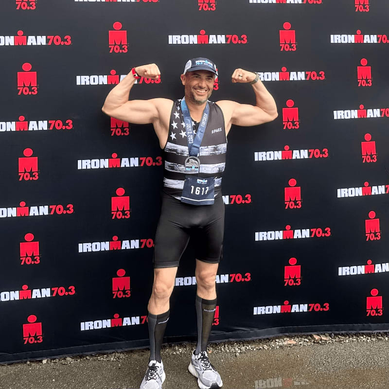 A muscular man in athletic gear flexes his biceps in the foreground against a black backdrop featuring repeated "IRONMAN 70.3" logos.