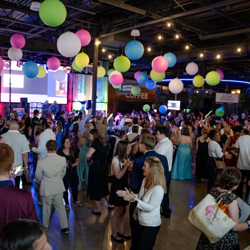 A crowd of people mingles in a large industrial venue decorated with colorful hanging lanterns and string lights.