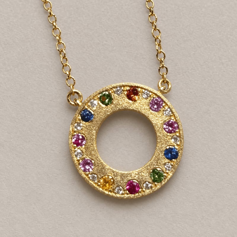 A gold circular pendant set with colorful gemstones hangs from a gold chain against a plain, light-grey background.