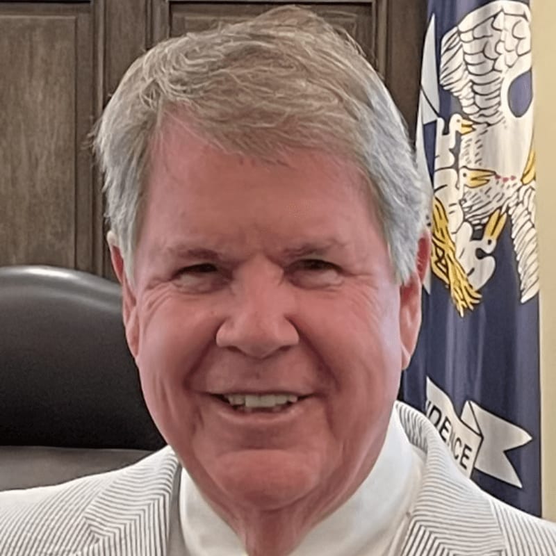 A smiling man in a light-colored striped blazer stands in the foreground against a background of a dark wooden wall and a blue flag.