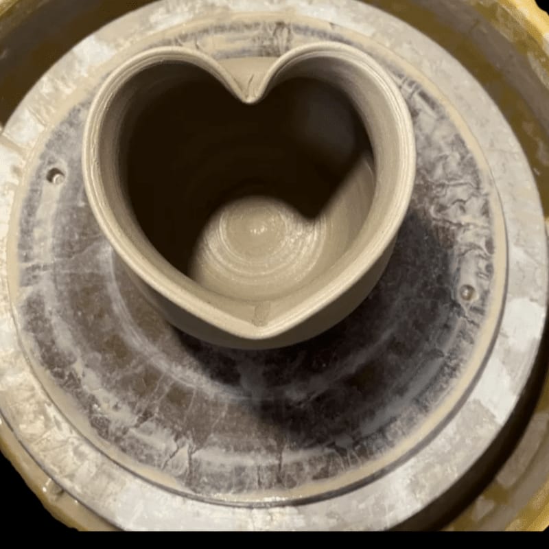 A heart-shaped ceramic pot sits on a potter's wheel, with the wheel's textured surface visible in the background.