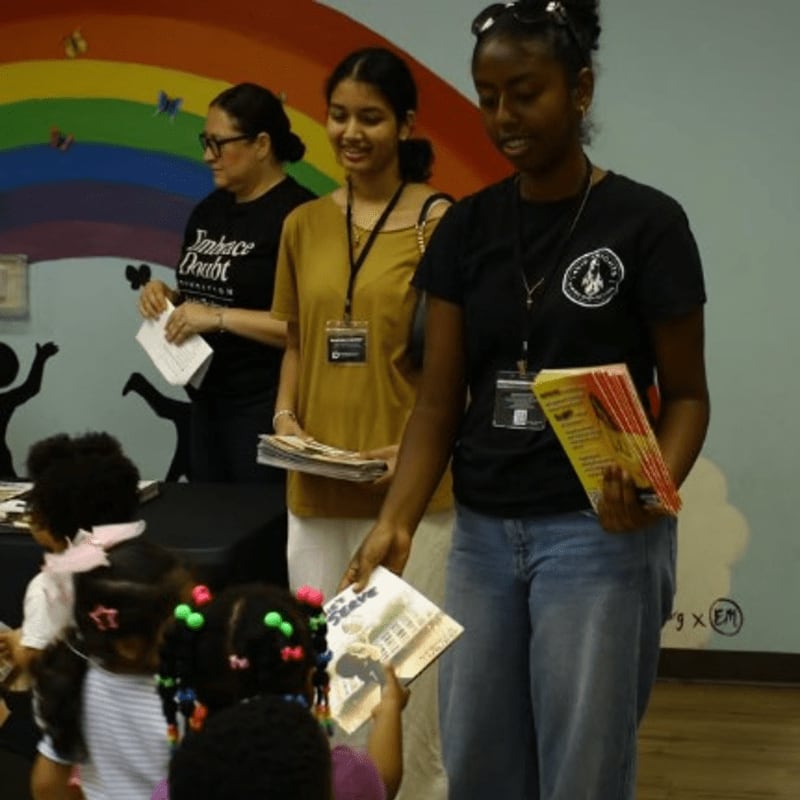 Three women stand in the foreground holding papers and books, with a colorful rainbow mural decorating the background wall.