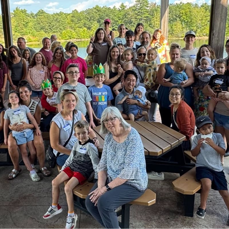 A large, multi-generational group of people poses for a photo around a wooden picnic table in the foreground, with a lush green forest and a lake in the background.