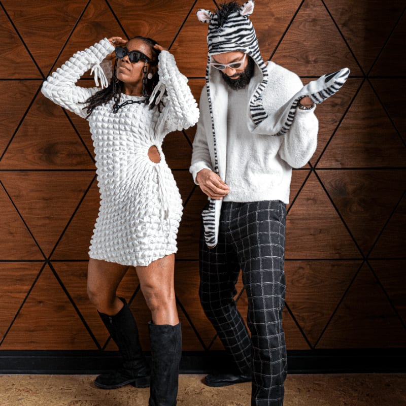 A woman in a white textured dress and a man in a zebra-themed hoodie and plaid pants pose against a dark wood-paneled wall.