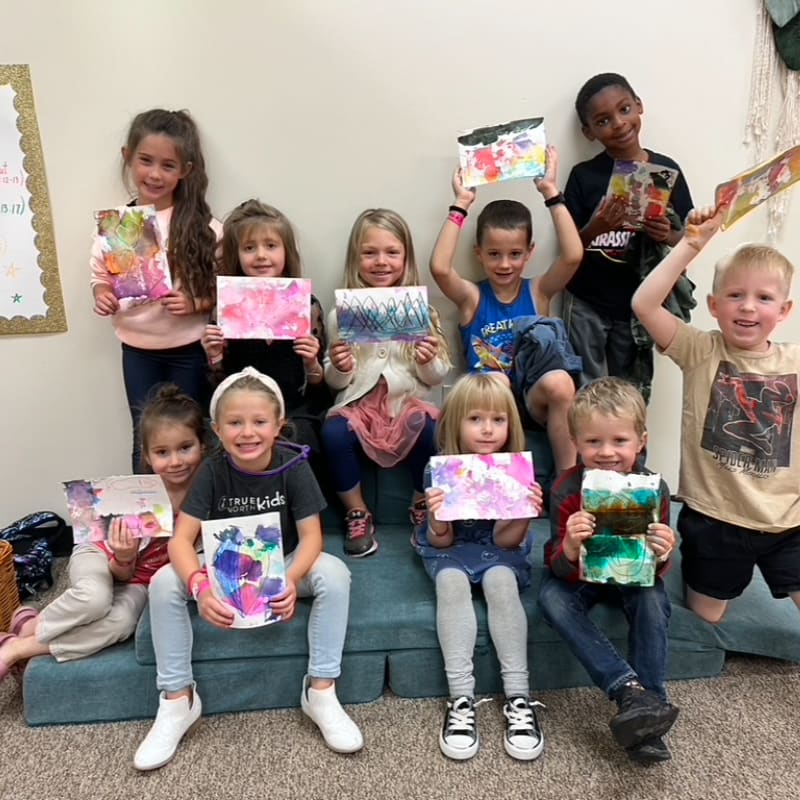 A group of smiling children hold up their colorful paintings while sitting and standing in a room with plain walls.