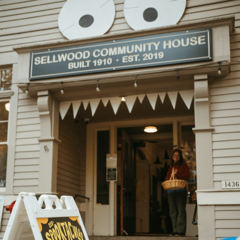 A white A-frame sign stands in the foreground before the entrance of the Sellwood Community House, which is decorated with large googly eyes and a jagged banner.
