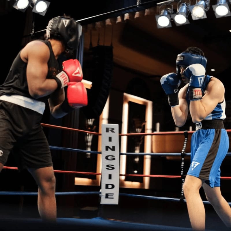 Two boxers wearing headgear and gloves face off in a boxing ring under bright overhead lights.