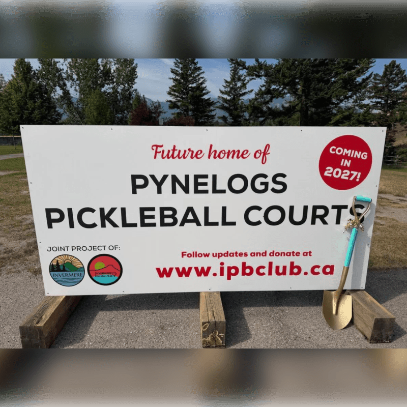 A white sign announcing the future home of Pynelogs Pickleball Courts stands in the foreground with a shovel leaning against it, set against a background of green trees and a clear sky.