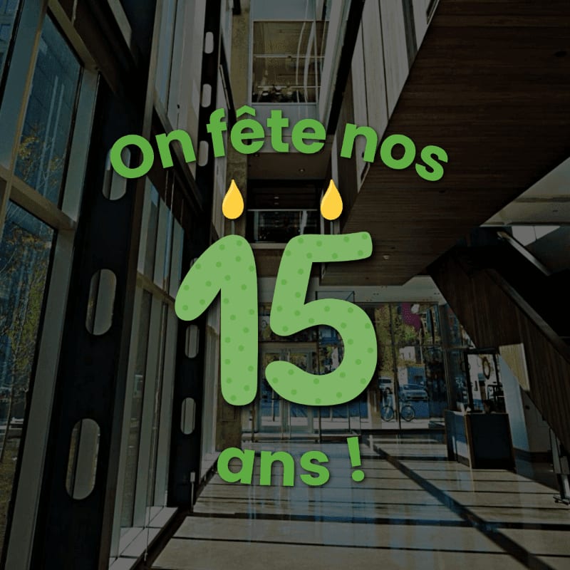 Green text reading "on fête nos 15 ans !" with two yellow droplets is overlaid on a modern building hallway with glass walls and a wooden ceiling.