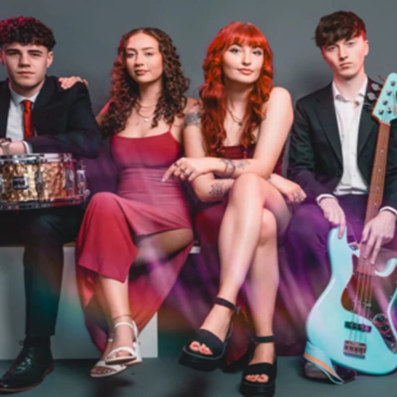 Four musicians pose together against a plain grey background, with a drummer and two women in red dresses sitting beside a guitarist.