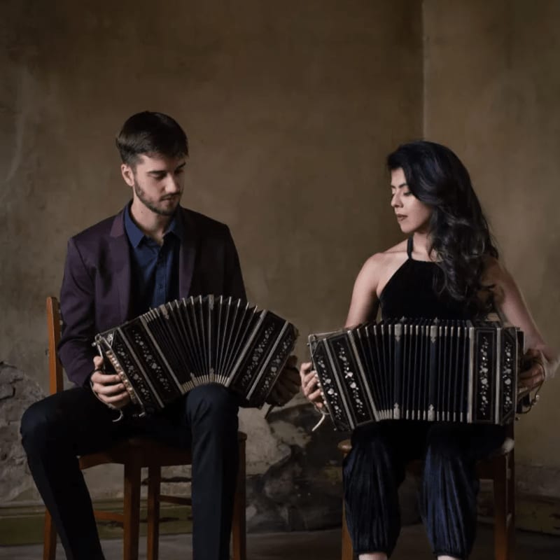 A man in a suit and a woman in a black dress sit on chairs playing accordions against a plain, neutral-toned wall.