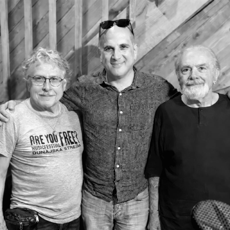 Three men stand side-by-side in the foreground against a background of light-colored wooden planks.