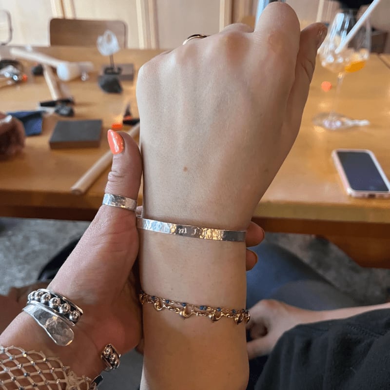 Two hands adorned with silver rings and bracelets are held together in the foreground, with a wooden table and a glass of wine in the blurred background.