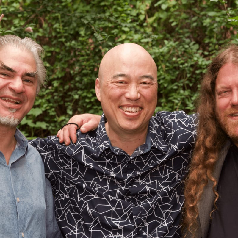 Three smiling men stand close together in the foreground against a lush background of green foliage.